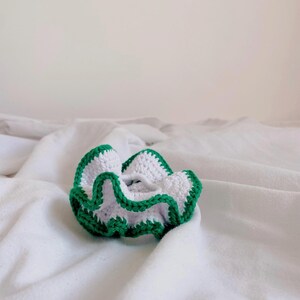 May include: A white crochet scrunchie with a green trim. The scrunchie is made of a soft, textured yarn and is designed to hold hair in place. The scrunchie is shown on a white background.