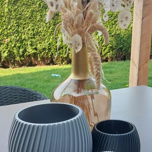 Op de afbeelding: Een decoratieve opstelling met een heldere glazen vaas met een gouden hals, gevuld met gedroogde bloemen en zaaddozen. Drie decoratieve, geribbelde, ronde plantenbakken in grijs, zwart en wit staan op de voorgrond.