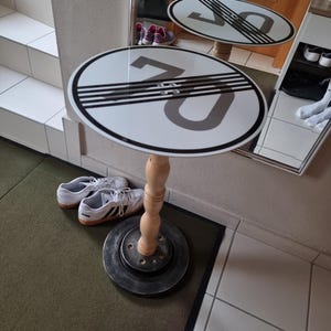 May include: A unique side table featuring a white tabletop with a black and gray "70" speed limit sign design. The table has a wooden spindle leg and a dark gray, circular base. A pair of white sneakers with black stripes are next to the table.