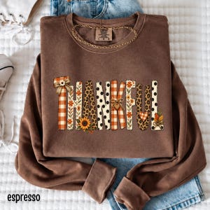 May include: A brown long-sleeve crewneck sweatshirt featuring the word THANKFUL printed in large letters. Each letter displays a unique pattern including orange gingham, floral prints, leopard spots, and black polka dots with small bows.