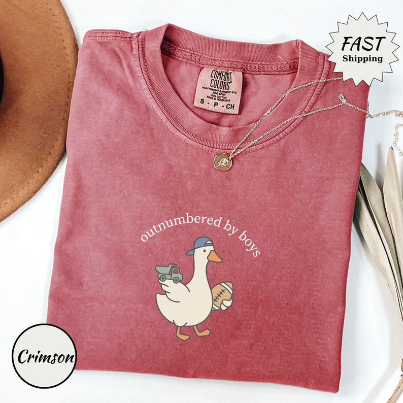 Op de afbeelding: Karmizijnrood T-shirt met de tekst "outnumbered by boys" en een cartoon eend met een baseballpet en een voetbal en een speelgoedvrachtwagen. Het shirt heeft een ronde hals en korte mouwen. Een gouden ketting is over het shirt gedrapeerd.