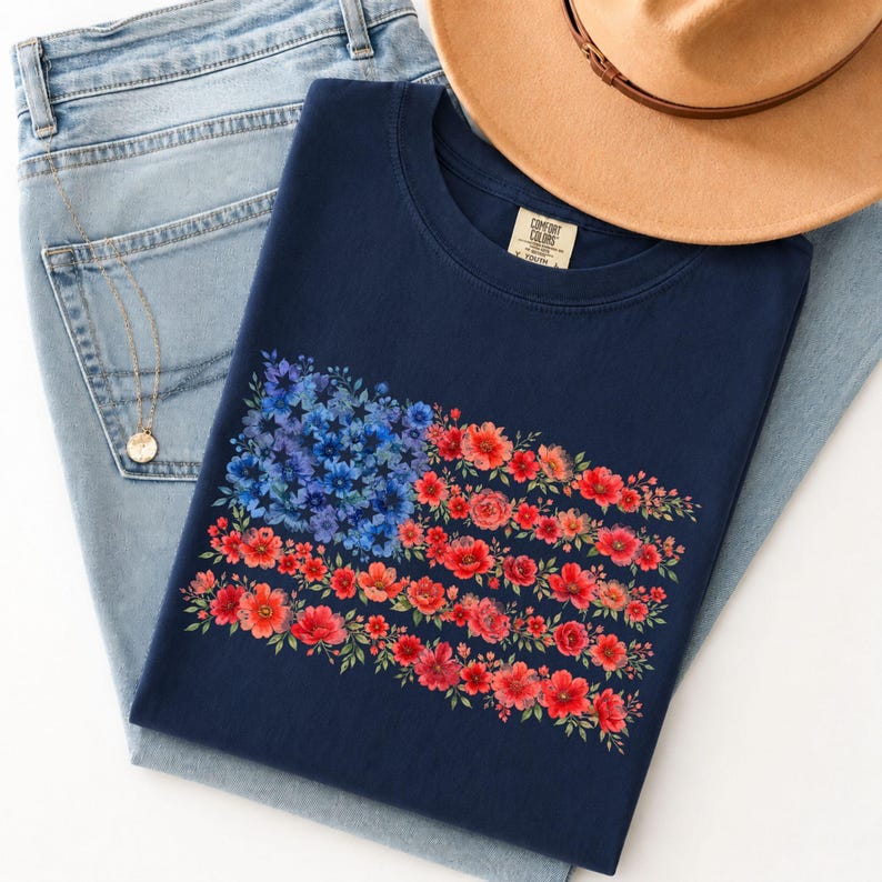 May include: A navy blue t-shirt featuring a floral American flag design. The flag is composed of blue flowers in the upper left corner and red and orange flowers forming the stripes. The shirt is laid on top of light blue jeans and a tan hat.