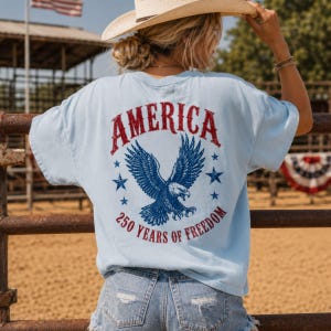 May include: Light blue t-shirt with the word "AMERICA" in red, a blue eagle graphic, and the text "250 YEARS OF FREEDOM." The shirt is made of a soft material and is suitable for casual wear.