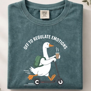 Puede incluir: Camiseta Comfort Colors verde azulado con un ganso blanco en un scooter, sosteniendo café y con una mochila. El texto "OFF TO REGULATE EMOTIONS" está sobre el ganso. Una pequeña etiqueta Comfort Colors es visible.