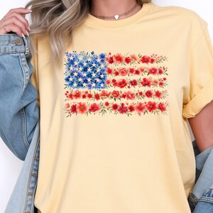 May include: A light yellow t-shirt featuring a floral American flag design. The flag's stars are composed of blue flowers, and the stripes are made of red flowers. The shirt has short sleeves and a crew neck.