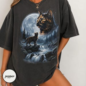 May include: A charcoal gray short-sleeve t-shirt featuring a graphic print of a tortoiseshell cat looking up at a large full moon. Below, a smaller cat stands on a rock near a flowing waterfall and pine trees. The word pepper is printed in black.