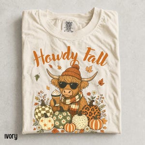 Può includere: Una t-shirt color avorio con la grafica di una mucca highland che indossa un berretto, occhiali da sole e una sciarpa mentre tiene una tazza di caffè. Il testo Howdy Fall è stampato sopra, circondato da zucche decorate e foglie autunnali.