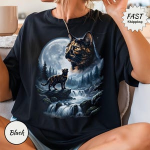 May include: A black t-shirt featuring a graphic print of a tortoiseshell cat looking toward a large full moon above a forest and a flowing waterfall. A smaller tortoiseshell cat stands on a rock in the foreground. The design includes a star-filled sky.