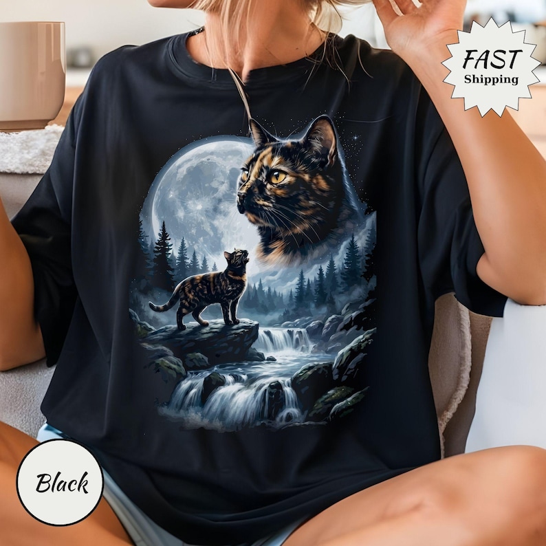 May include: A black t-shirt featuring a graphic print of a tortoiseshell cat looking toward a large full moon above a forest and a flowing waterfall. A smaller tortoiseshell cat stands on a rock in the foreground. The design includes a star-filled sky.