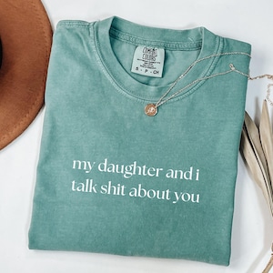 May include: A seafoam green t-shirt with the text "my daughter and i talk shit about you" in white. The shirt has a crew neck and short sleeves. A silver necklace with a small pendant is draped across the shirt.