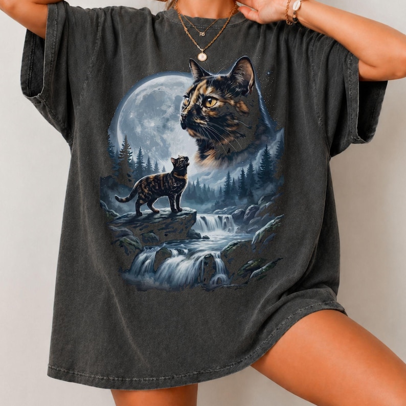 May include: A charcoal gray short-sleeve t-shirt featuring a graphic print of a large cat face in the sky above a smaller cat standing on a rock near a waterfall, a forest, and a full moon. The shirt has a crew neckline and a relaxed fit.