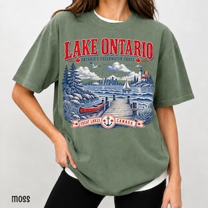 K&ouml;nnte beinhalten: Ein moosgr&uuml;nes Kurzarm-T-Shirt mit einem Grafikdruck des Lake Ontario. Das Design zeigt ein rotes Kanu, einen Holzsteg, einen Leuchtturm und eine Stadtsilhouette. Der Text lautet LAKE ONTARIO, ONTARIO'S FRESHWATER COAST und GREAT LAKES CANADA.