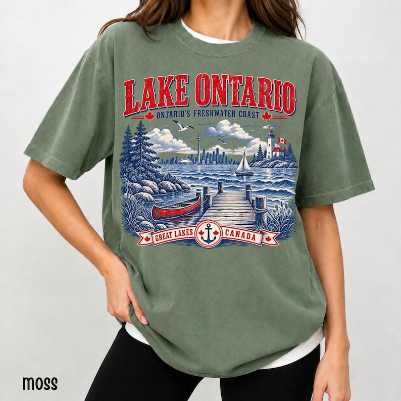 K&ouml;nnte beinhalten: Ein moosgr&uuml;nes Kurzarm-T-Shirt mit einem Grafikdruck des Lake Ontario. Das Design zeigt ein rotes Kanu, einen Holzsteg, einen Leuchtturm und eine Stadtsilhouette. Der Text lautet LAKE ONTARIO, ONTARIO'S FRESHWATER COAST und GREAT LAKES CANADA.