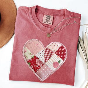 Peut inclure: Un t-shirt rose poudré Comfort Colors avec un motif de cœur en patchwork. Le cœur est composé de divers tissus à motifs roses et rouges, notamment des motifs floraux, à pois et vichy. Un délicat collier en argent avec un petit pendentif repose sur le t-shirt.