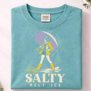 May include: A turquoise Comfort Colors t-shirt featuring a graphic of a person walking in the rain under a purple umbrella. The person is wearing a yellow jacket and boots. The text "stay SALTY melt ICE" is printed below the graphic.