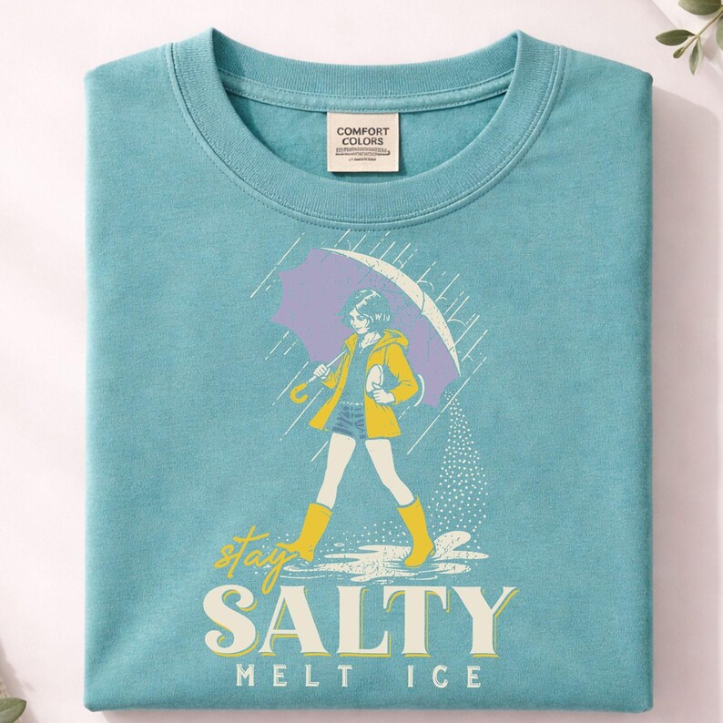 May include: A turquoise Comfort Colors t-shirt featuring a graphic of a person walking in the rain under a purple umbrella. The person is wearing a yellow jacket and boots. The text "stay SALTY melt ICE" is printed below the graphic.