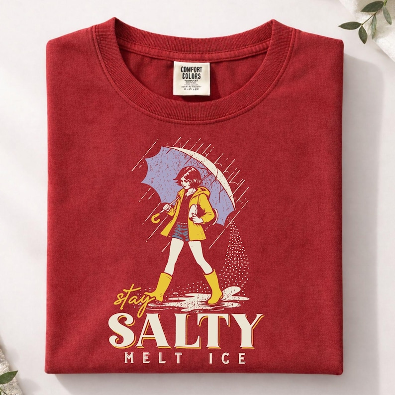 May include: A red Comfort Colors t-shirt featuring an illustration of a person in a yellow raincoat and boots holding a blue umbrella in the rain. The text "stay SALTY melt ICE" is below the illustration.