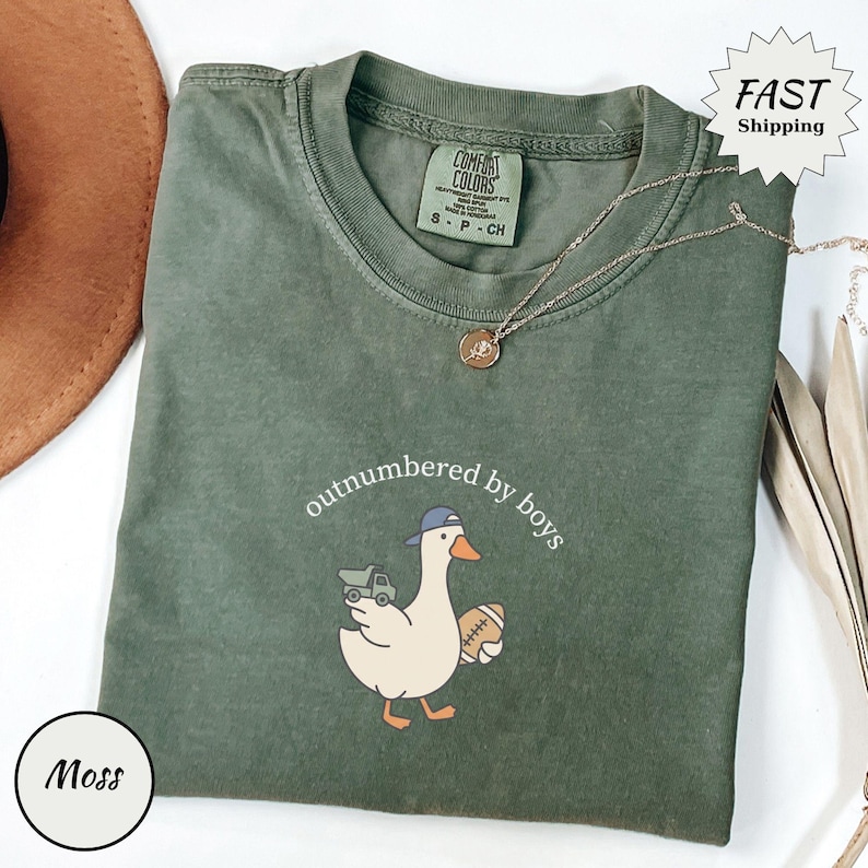 Op de afbeelding: Mosgroen t-shirt met de tekst "outnumbered by boys" en een cartoon eend met een baseballpet en een voetbal. Het shirt heeft een Comfort Colors label en een gouden ketting.