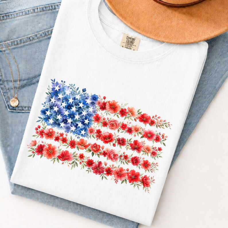 May include: White t-shirt featuring a floral American flag design. The flag is composed of blue and white flowers representing the stars and red flowers for the stripes. The shirt is folded on a pair of blue jeans.