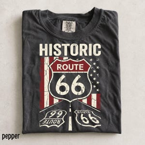 以下が含まれることがあります： チャコールグレーの半袖Tシャツ。中央にアメリカ国旗を様式化したデザインと、かすれた質感の「Historic Route 66」の盾のグラフィックがプリントされています。下部には小さな盾のアイコンと「Route 66」の文字が配置されています。