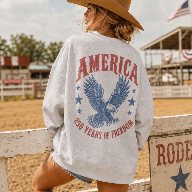 May include: A light gray sweatshirt with the word "AMERICA" in red, a blue eagle graphic, and the text "250 YEARS OF FREEDOM." The sweatshirt is worn by a person wearing a brown cowboy hat.
