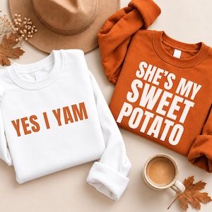 Puede incluir: Una sudadera blanca con el texto naranja YES I YAM y una sudadera color naranja quemado con el texto blanco SHE'S MY SWEET POTATO. Las prendas se muestran junto a un sombrero marrón, flores secas, hojas de otoño y una taza de café.