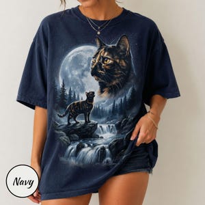May include: A navy blue short-sleeve t-shirt featuring a graphic print of a tortoiseshell cat looking toward a large full moon. Below, a smaller cat stands on a rocky cliff overlooking a waterfall surrounded by a dark forest of pine trees.