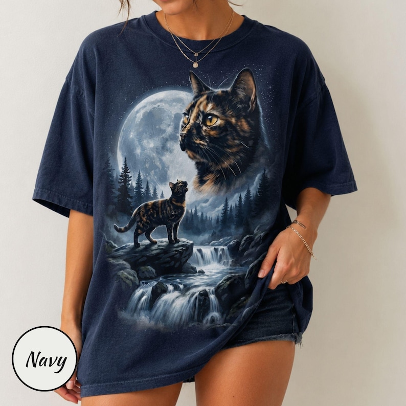 May include: A navy blue short-sleeve t-shirt featuring a graphic print of a tortoiseshell cat looking toward a large full moon. Below, a smaller cat stands on a rocky cliff overlooking a waterfall surrounded by a dark forest of pine trees.