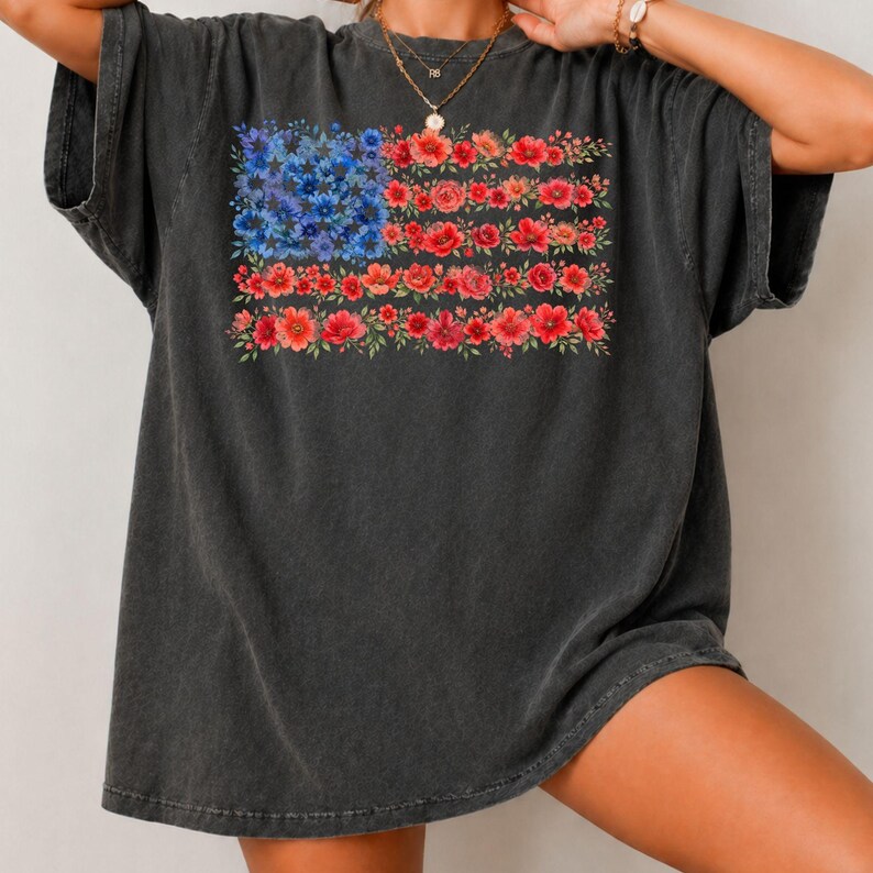 May include: Dark gray t-shirt featuring a floral American flag design. The flag's stars are represented by blue flowers, and the stripes are made of red flowers. The shirt has short sleeves and a relaxed fit.