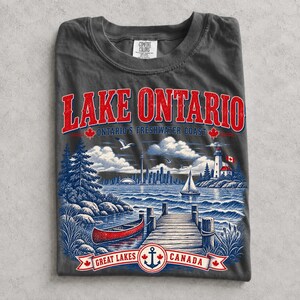 K&ouml;nnte beinhalten: Ein anthrazitfarbenes T-Shirt mit einem Grafikdruck des Lake Ontario. Das Design zeigt eine rote Text&uuml;berschrift, eine blaue Landschaft mit einem Holzsteg, einem roten Kanu, einem Segelboot, einem Leuchtturm und einer Skyline sowie ein Banner mit Great Lakes Canada.