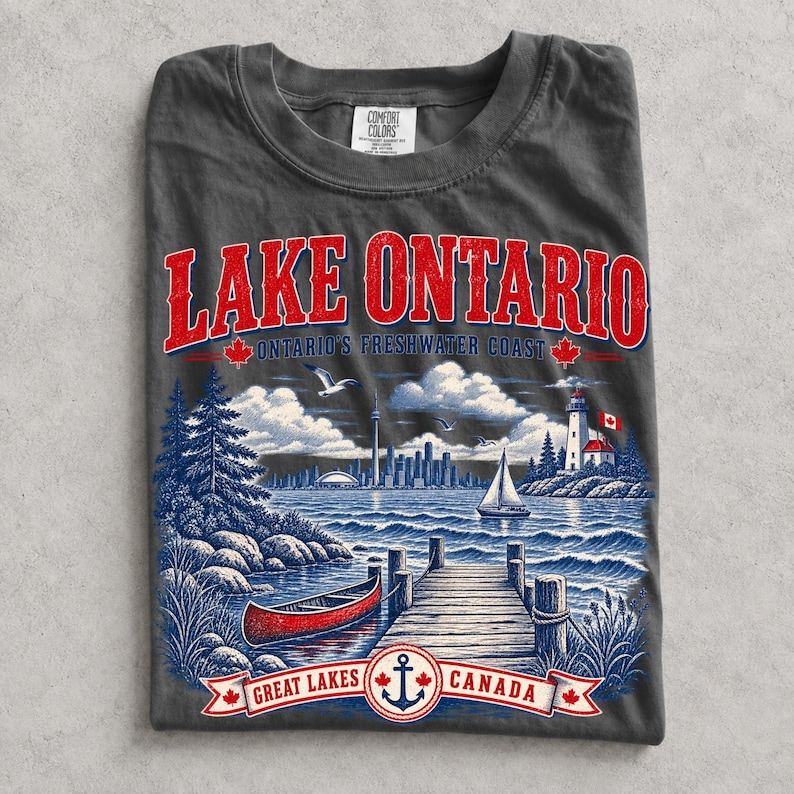 K&ouml;nnte beinhalten: Ein anthrazitfarbenes T-Shirt mit einem Grafikdruck des Lake Ontario. Das Design zeigt eine rote Text&uuml;berschrift, eine blaue Landschaft mit einem Holzsteg, einem roten Kanu, einem Segelboot, einem Leuchtturm und einer Skyline sowie ein Banner mit Great Lakes Canada.