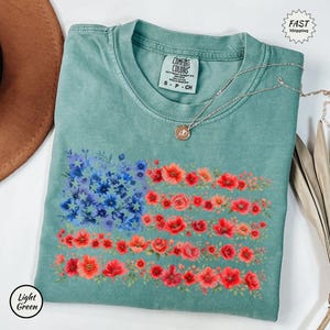 May include: A light green t-shirt featuring a floral American flag design. The flag is composed of blue and red flowers, with a silver necklace draped across the shirt. The shirt has a Comfort Colors tag and the words "Light Green" are in a circle.