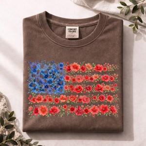 May include: Brown t-shirt featuring a floral American flag design. The flag is composed of blue and red flowers, with the text "COMFORT COLORS" on the tag. The shirt is folded and displayed on a neutral surface.