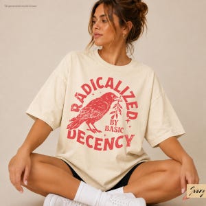 May include: An ivory short-sleeved t-shirt featuring a distressed red graphic of a crow holding a branch. The text RADICALIZED BY BASIC DECENCY is printed in a circular red font around the bird. The shirt has a loose, relaxed fit.