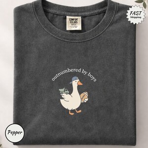 Op de afbeelding: Donkergrijs Comfort Colors T-shirt met een afbeelding van een gans met een blauwe pet en een voetbal en een speelgoedvrachtwagen. De tekst "outnumbered by boys" staat boven de gans. Het shirt is opgevouwen.