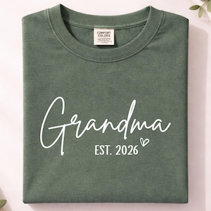May include: An olive green t-shirt, folded neatly, featuring the word "Grandma" in white script. Underneath, the text reads "EST. 2026" with a small heart. The shirt has a crew neck and a "Comfort Colors" label.