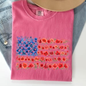 May include: A pink t-shirt featuring a floral American flag design. The flag's stars are blue flowers, and the stripes are composed of red and orange blooms. The shirt is folded, with the Comfort Colors label visible.