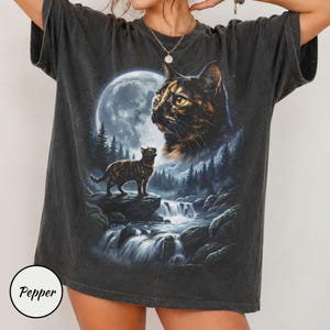 Puede incluir: Una camiseta de manga corta gris carbón con un estampado gráfico de un gato carey de pie sobre una roca cerca de una cascada, con una luna grande y una segunda cara de gato más grande al fondo. La palabra Pepper está impresa en un círculo.