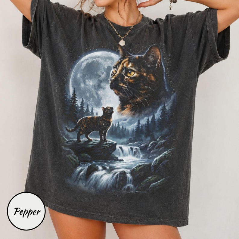 May include: A charcoal gray short-sleeve t-shirt featuring a graphic print of a tortoiseshell cat standing on a rock near a waterfall, with a large moon and a second, larger cat face in the background. The word Pepper is printed in a small circle.