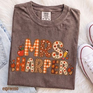 Può includere: Una t-shirt marrone a maniche corte con scollo rotondo ed etichetta Comfort Colors. La grafica presenta la scritta MRS HARPER in un font a motivi misti con stampe a quadretti arancioni, floreali e foglie, accentuata da una mela rossa e una matita.