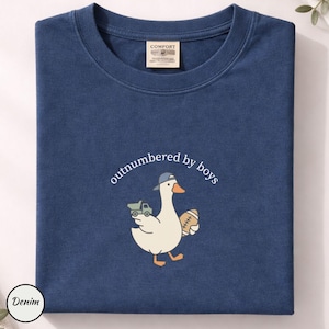 Op de afbeelding: Marineblauwe t-shirt met een ronde hals. De shirt heeft een cartoon gans met een baseballpet en houdt een voetbal en een speelgoedvrachtwagen vast. De tekst "outnumbered by boys" staat boven de gans.