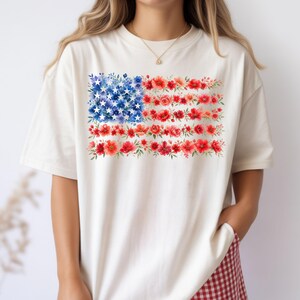 May include: Cream-colored t-shirt featuring a floral American flag design. The flag's stars are represented by blue flowers, and the stripes are formed by red flowers. The shirt has short sleeves and a relaxed fit.