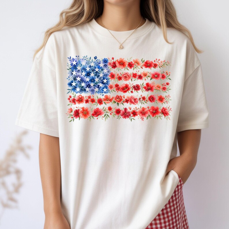 May include: Cream-colored t-shirt featuring a floral American flag design. The flag's stars are represented by blue flowers, and the stripes are formed by red flowers. The shirt has short sleeves and a relaxed fit.
