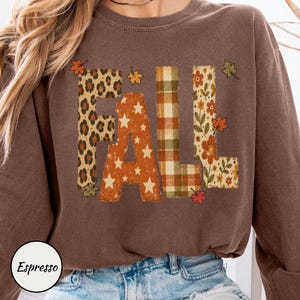 May include: A brown crewneck sweatshirt featuring the word FALL printed in large block letters. Each letter has a unique pattern: leopard print, orange with white stars, green and tan plaid, and a floral design with scattered autumn leaves.