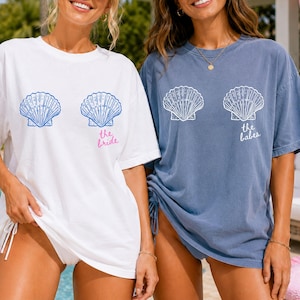 May include: A white t-shirt with blue seashell graphics and the pink text the bride, and a blue t-shirt with white seashell graphics and the white text the babes. Both shirts feature short sleeves and a crew neckline in a relaxed fit.