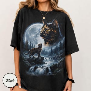 May include: A black short-sleeve t-shirt featuring a graphic print of a tortoiseshell cat looking up at a large full moon. Below, a smaller cat stands on a rock near a flowing waterfall surrounded by a dark forest of pine trees.