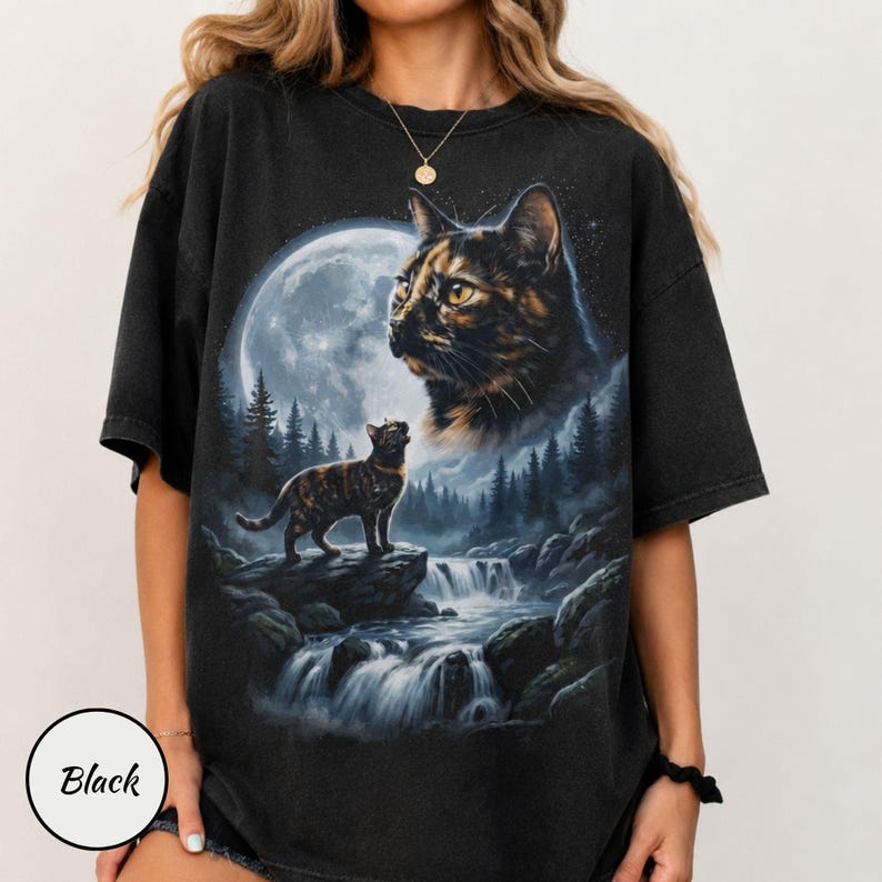 May include: A black short-sleeve t-shirt featuring a graphic print of a tortoiseshell cat looking up at a large full moon. Below, a smaller cat stands on a rock near a flowing waterfall surrounded by a dark forest of pine trees.