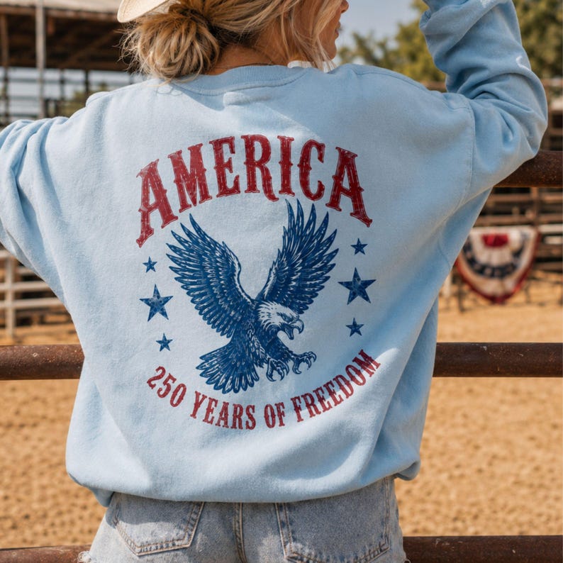 May include: Light blue sweatshirt with the word "AMERICA" in red, a blue eagle graphic, and the text "250 YEARS OF FREEDOM." The design includes stars. The sweatshirt is a casual style.
