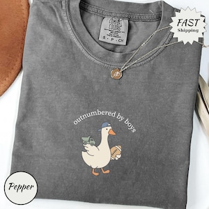 Op de afbeelding: Een grijs Comfort Colors T-shirt met de tekst "outnumbered by boys" en een cartoon eend met een baseballpet en een voetbal en een speelgoedvrachtwagen. Een gouden ketting is over het shirt gedrapeerd.