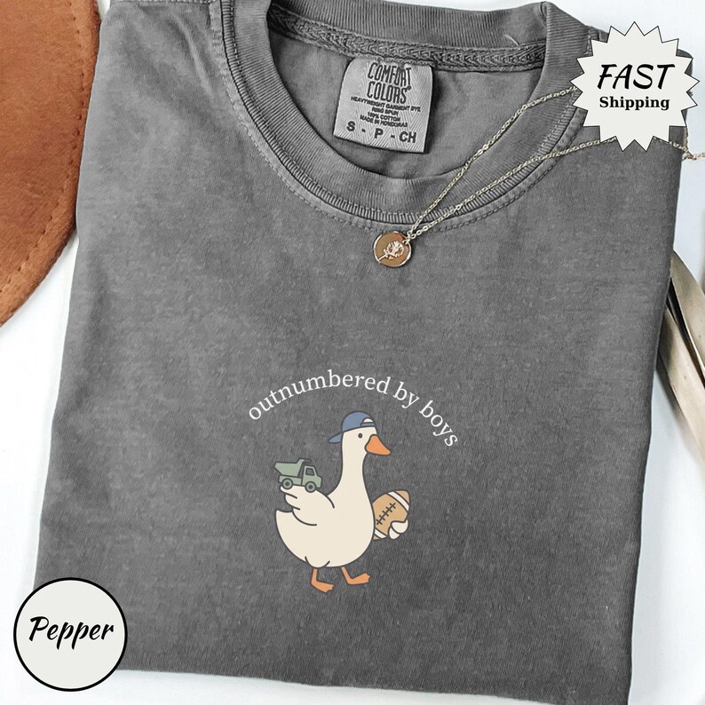 Op de afbeelding: Een grijs Comfort Colors T-shirt met de tekst "outnumbered by boys" en een cartoon eend met een baseballpet en een voetbal en een speelgoedvrachtwagen. Een gouden ketting is over het shirt gedrapeerd.